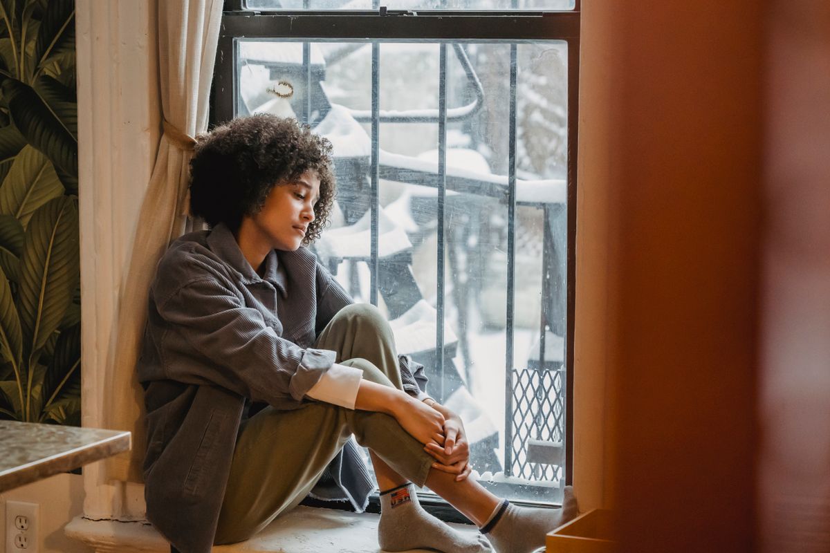 Femme repliée sur elle-même près d'une fenêtre, effets de la solitude sur la santé