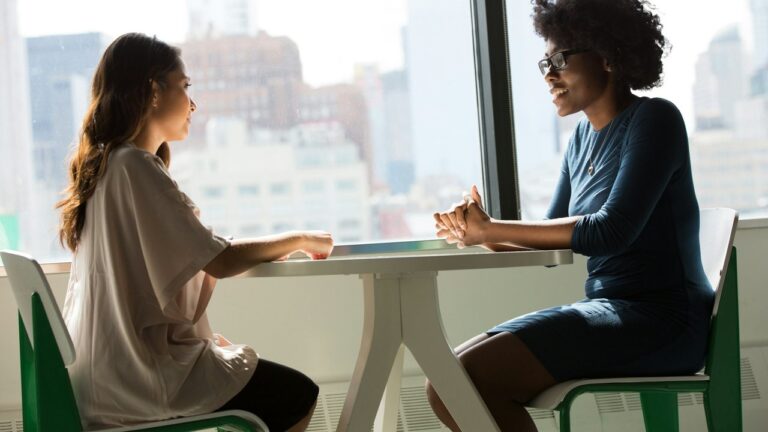 العنف الزوجي: امرأة تبوح لامرأة أخرى تنصت إليها، وهما جالستان وجهاً لوجه قرب نافذة