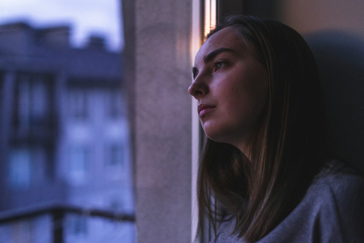 امرأة شابة متأملة تنظر من النافذة في ضوء المساء