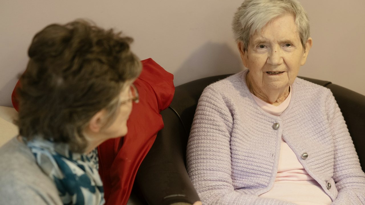 Alzheimer : une femme s'entretient calmement avec une dame âgée assise dans un fauteuil, chez elle