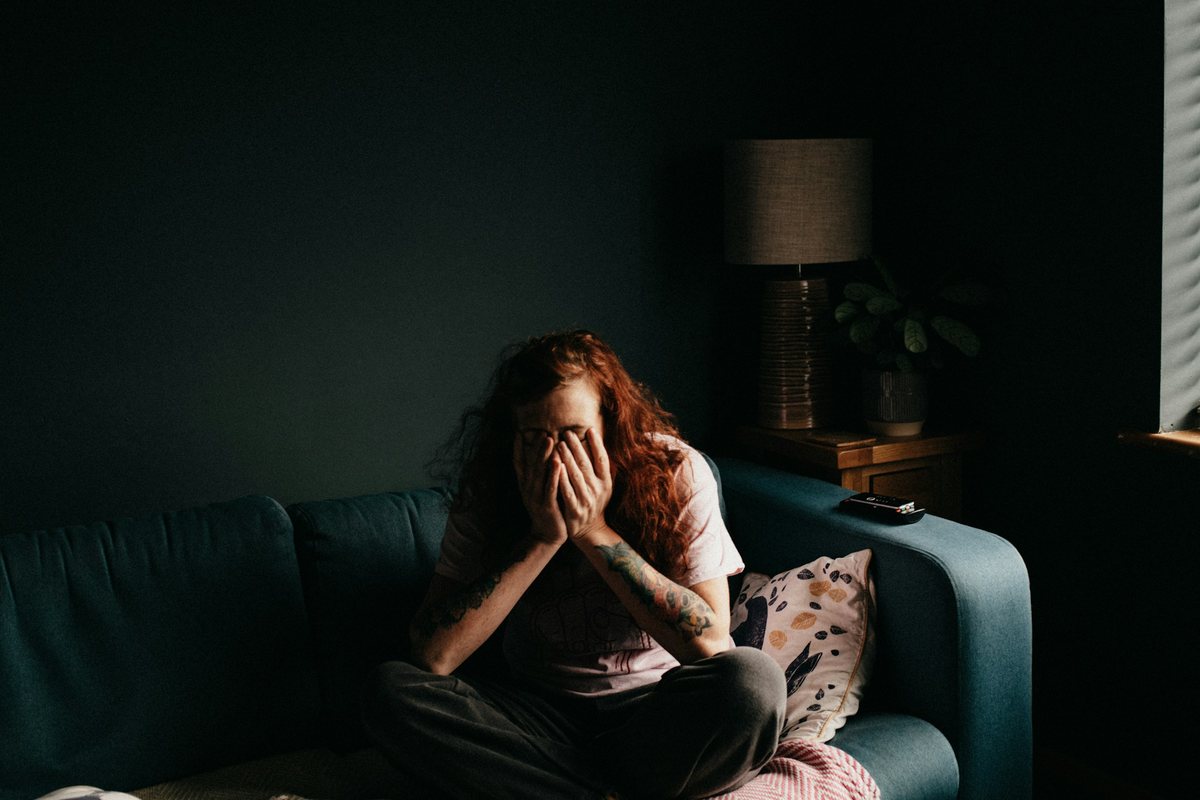 Symptômes psychiques du syndrome prémenstruel Femme assise sur un canapé dans la pénombre, le visage dans les mains : tristesse et irritabilité du syndrome prémenstruel